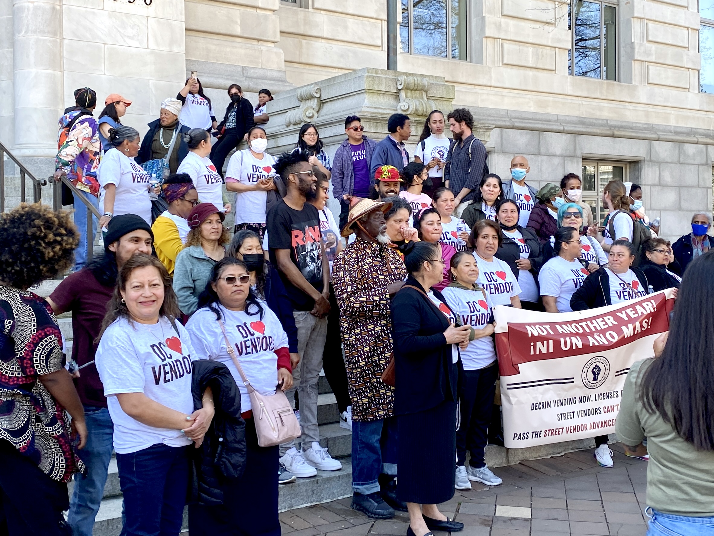 A large group of vendors, many in shirts that read vendadores unidos with a heart, stand on the steps of the Wilson Building. They appear to be cheering.