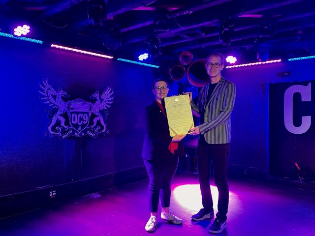 Photo of Councilmember Brianne K Nadeau presenting ceremonial resolution to Bill Spieler from DC9 Nightclub on stage at the venue. DC9 logo in background of photo.