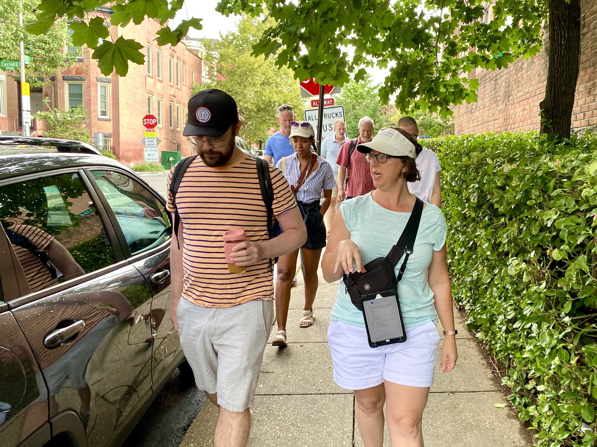 A large group of people is walking down the sidewalk toward the camera. In front is a man on the left with a coffee cup wearing shorts and a horizontal striped shirt. He's speaking to Councilmember Nadeau, also wearing shorts and a t-shirt and a white visor.