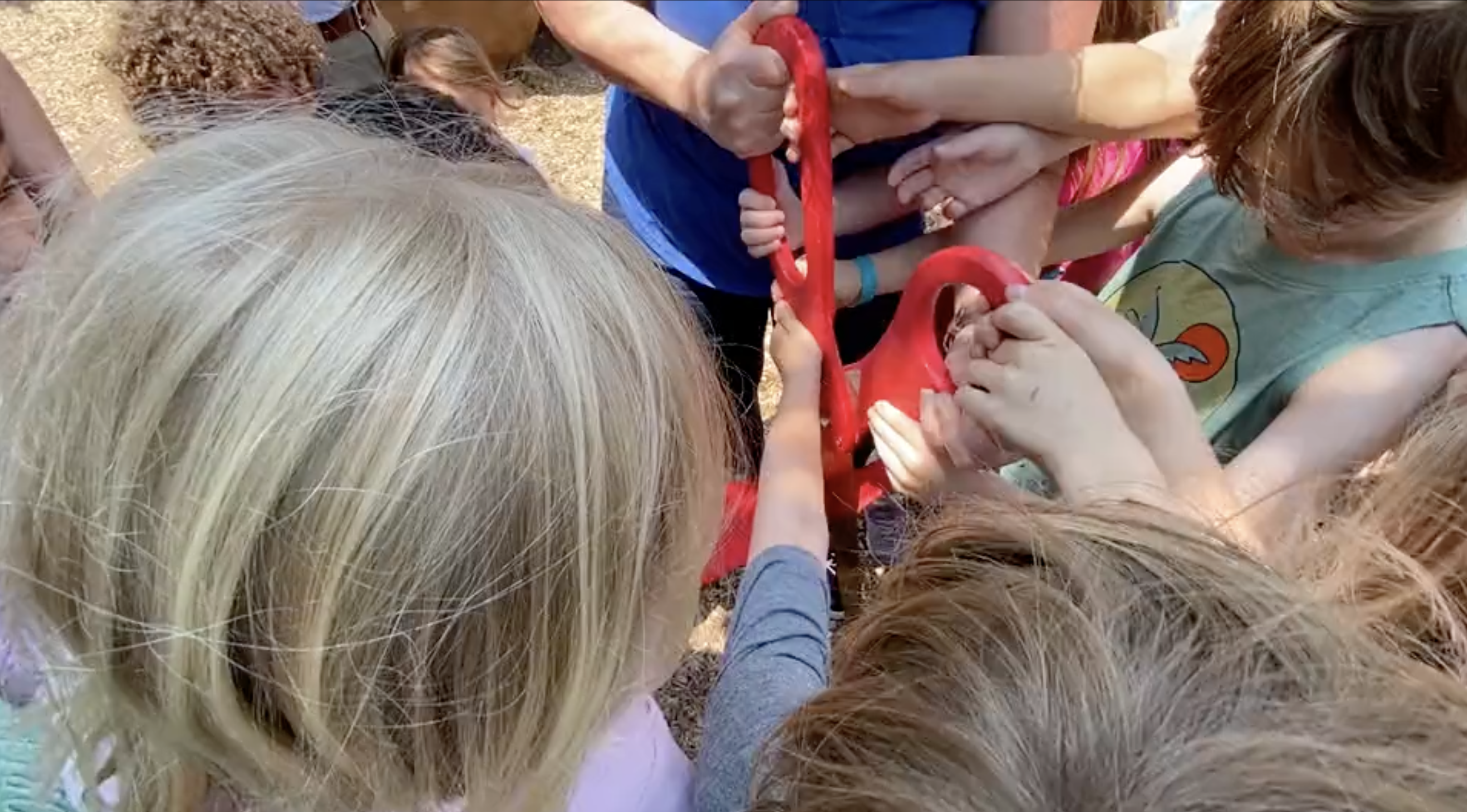 Close up from above of three young kids plus an adult holding oversized scissors with big red handles to cut a large ribbon.