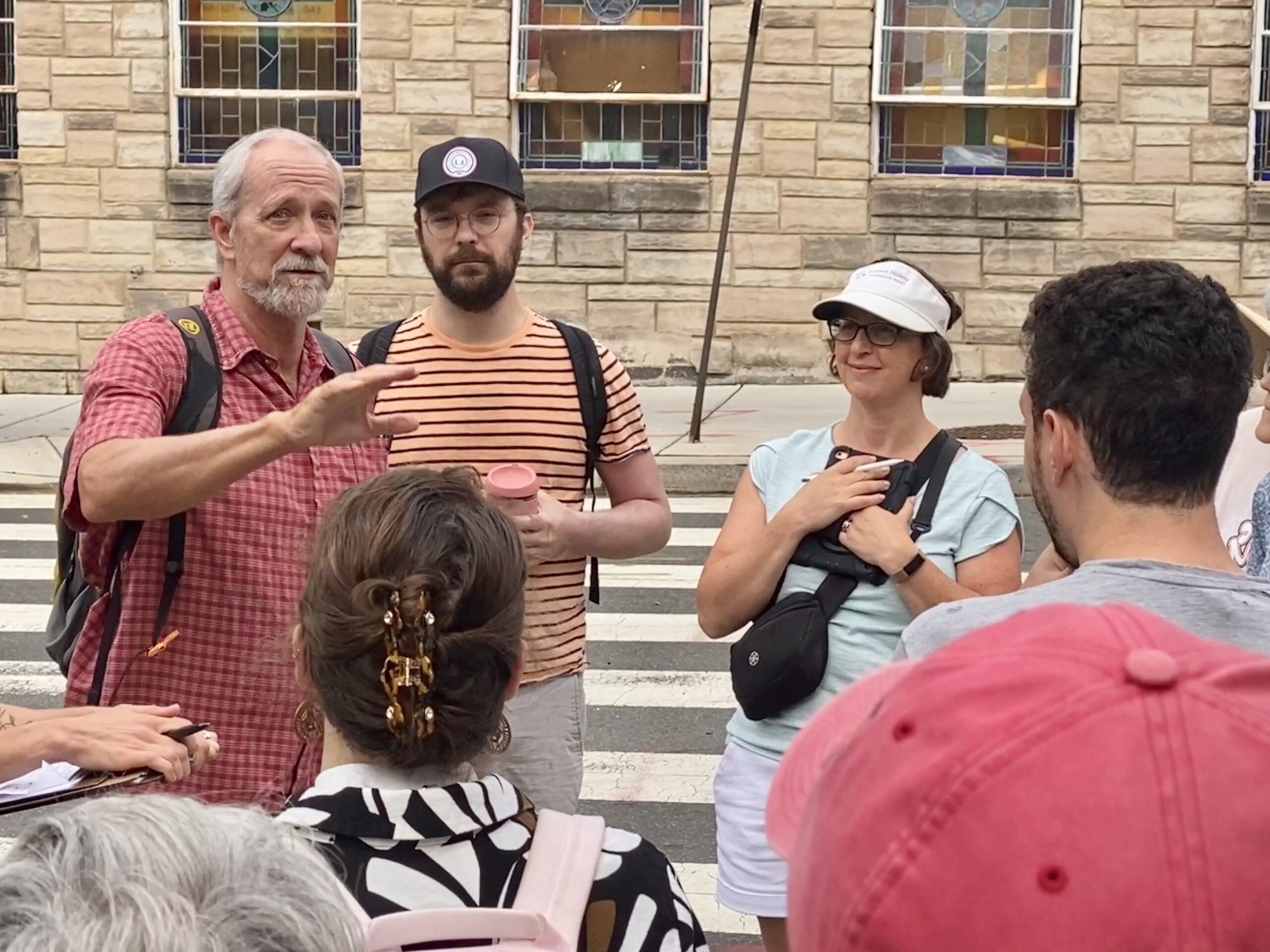 Man speaks to a group of people on the sidewalk at an intersection with Councilmember Nadeau standing next to him.