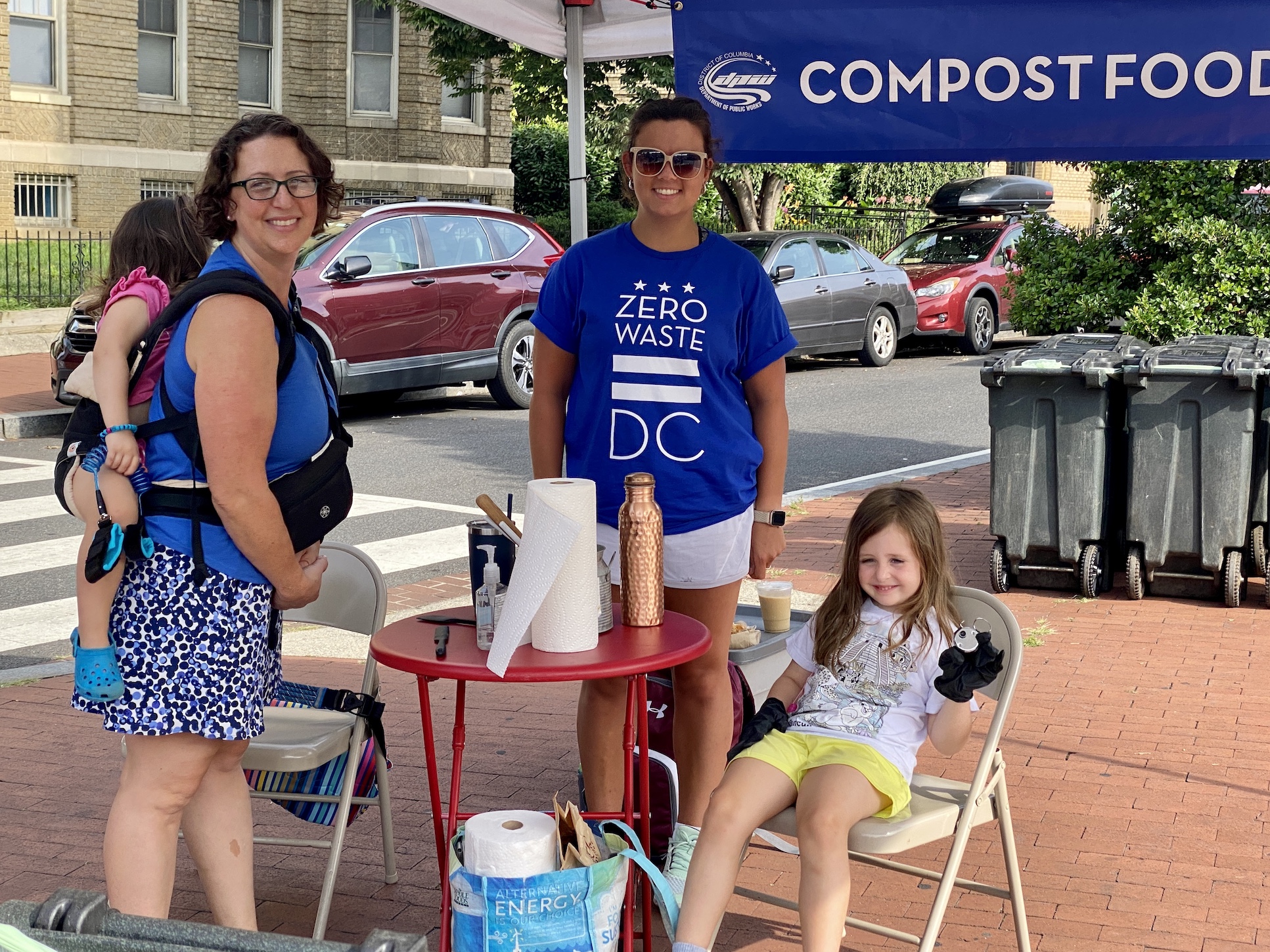 Councilmember Nadeau with child in a carrier on her back and a worker under a tent with a sign that reads Compost Food