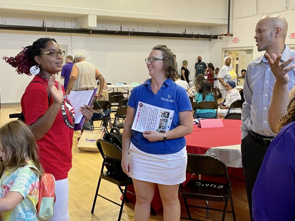 Three people standing, talking and smiling in a room full of tables and folding chairs and people - looks like a gymnasium at a school. Woman on the left in a red printed t-shirt and glasses gestures with her hand. Councilmember Nadeau, center, smiles and listens, while a man to the right with hands in his pocket looks and listens on.