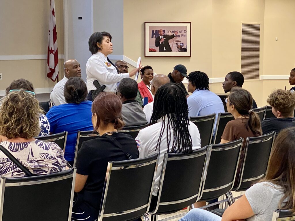 MPD Assistant Chief Morgan Kane at front of room speaking and gesturing to an audience. Behind her are four additional guest speakers facing the audience.