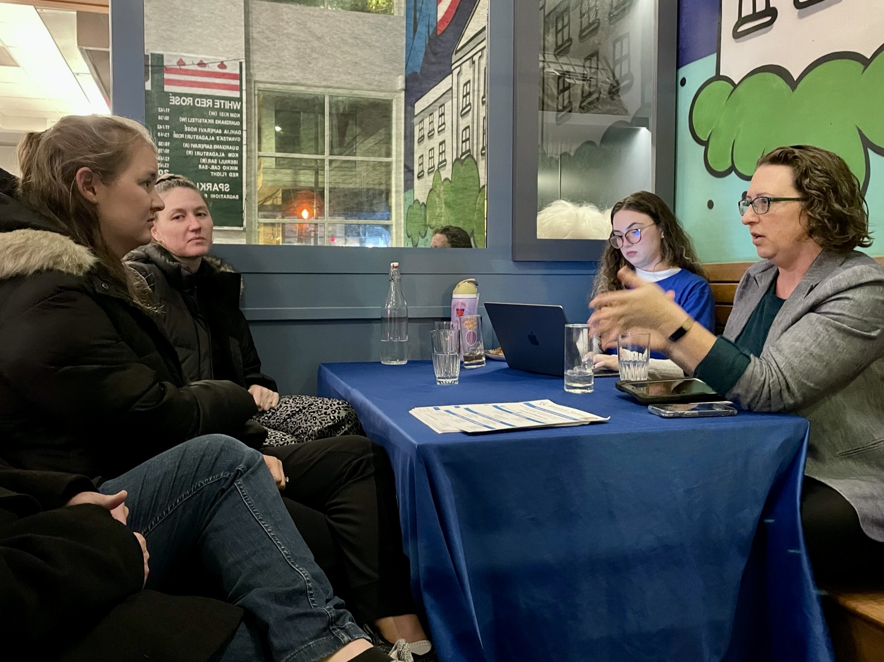 Councilmember Nadeau gestures as she speaks with two people sitting across the table from her. They are seated at a table in a restaurant. A staff person wearing a blue Brianne Nadeau t-shirt takes notes on a laptop.