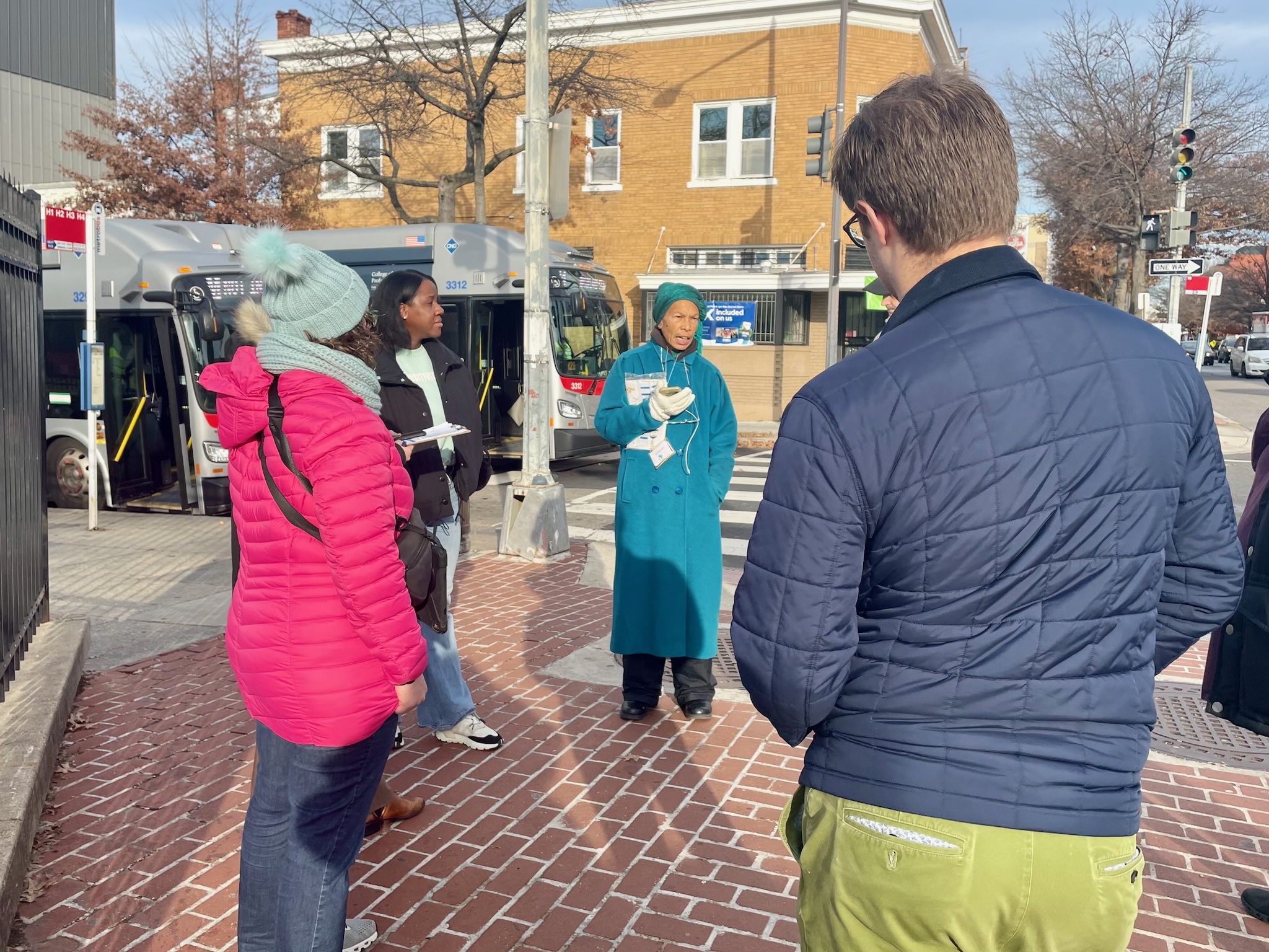 Councilmember Nadeau, back to camera, listening as a resident speaks. They and three others are on a sidewalk at a street corner. They are wearing winter coats.