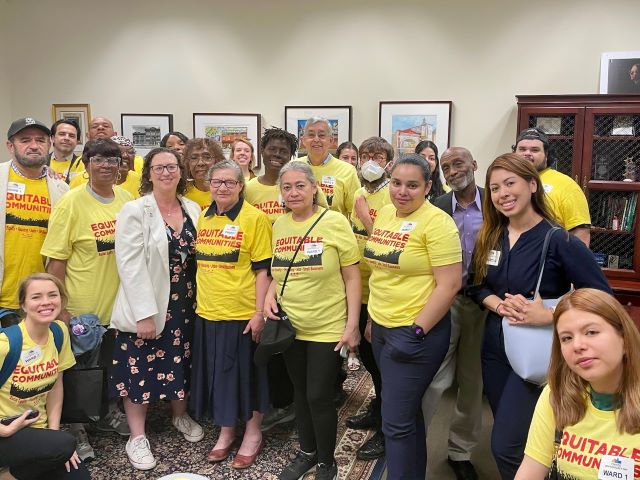 A group of people with yellow t-shirts that say Equitable Communities in red print gather with Councilmember Brianne K Nadeau in her office