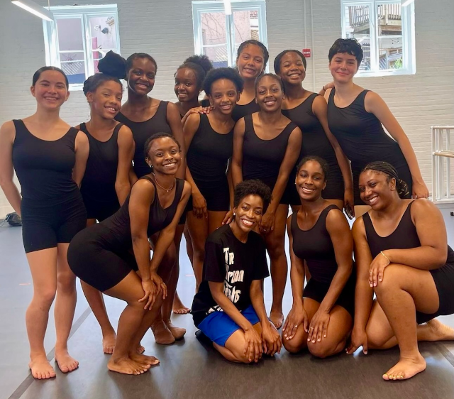 Thirteen teenage girls wearing black leotards pose for a photo in a dance rehearsal space