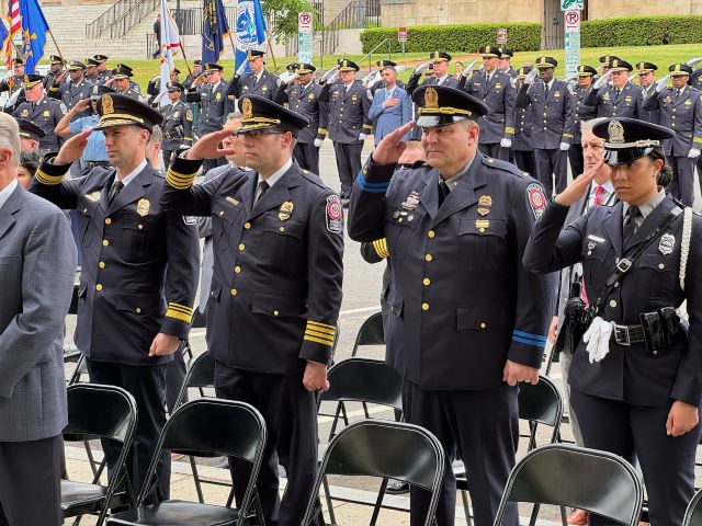 Councilmember Nadeau speaks with MPD Chief Pamela Smith before the ceremony begins; Officers carrying flags and ceremonial rifles march in front of a line of officers in uniform standing at attention; Officers in dress uniform stand and salute. Behind them a row of many other officers do the same. 