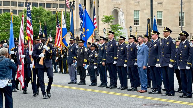 Councilmember Nadeau speaks with MPD Chief Pamela Smith before the ceremony begins; Officers carrying flags and ceremonial rifles march in front of a line of officers in uniform standing at attention; Officers in dress uniform stand and salute. Behind them a row of many other officers do the same. 