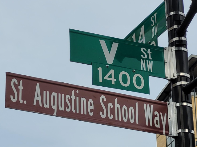 Brown street sign reads “St. Augustine School Way” below sign for V St. NW and one for 14th St. NW