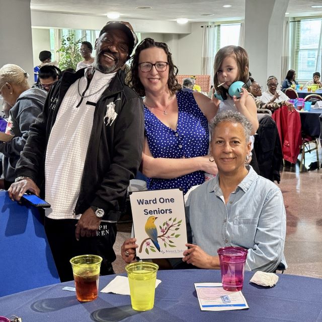 Photo of Councilmember Brianne K Nadeau holding her daughter and posing with two seniors seated at table at the Ward 1 Senior Dance