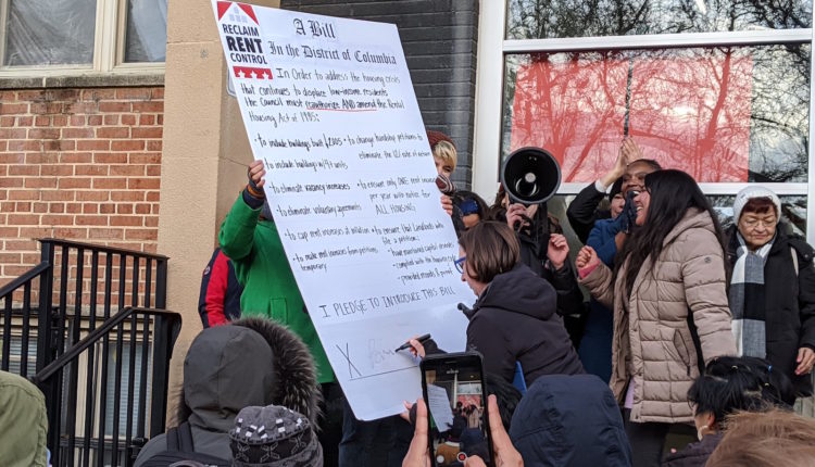 Councilmember Nadeau signs a "Reclaim Rent Control" pledge on an oversized posterboard being held up by a group of participants in an outdoor rally. A person stands nearby with a bullhorn.