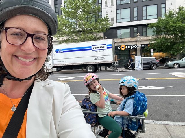 Councilmember Brianne K Nadeau smiles in her bike helmet with her two daughters on the back of her bike on Bike to Work Day