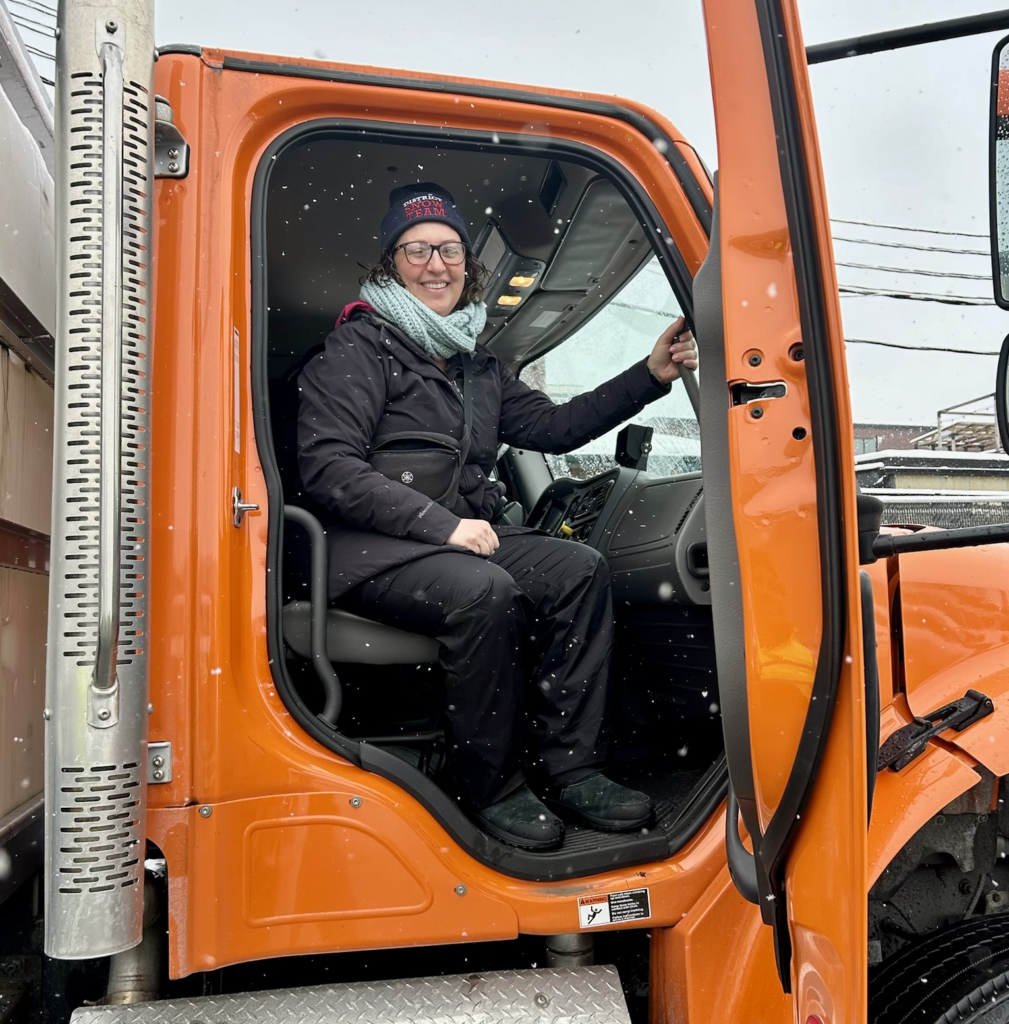 Councilmember Nadeau sits in passernger side of the cab of a large plow truck with orange door. The door is open and she is looking out and smiling.