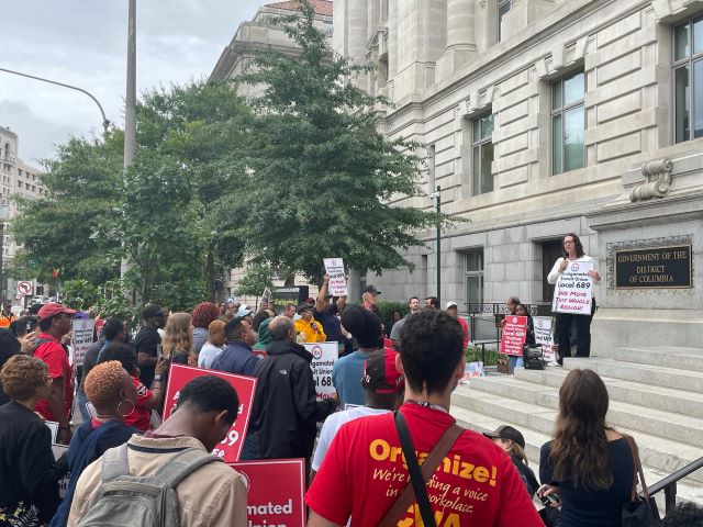 Councilmember Brianne K Nadeau speaks from the Wilson Building steps to a group of people holding ATU rally signs. Nadeau holds a sign as well.