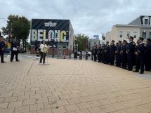 Councilmember Brianne K Nadeau speaks at a podium with DC Fire logo on front. A line of firefighters on the right hand side stand listening.