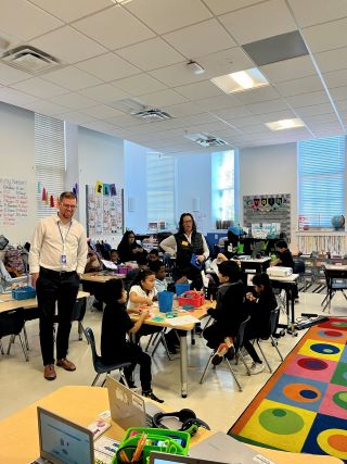 Teacher and Councilmember Brianne K Nadeau stand in classroom with students sitting at round tables. Colorful rug is in right of photo.