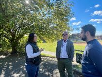 Councilmember Brianne K Nadeau speaks to Councilmember Matt Frumin and a male staffer at KC Lewis in front of a tree with sun streaming through its branches.