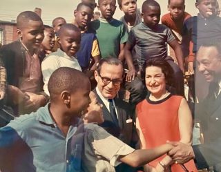 Young children surround Lady Bird Johnson, pictured center smiling in a red dress. One child shakes the hand of a man in a black suit and tie on the right, while another man in a black suit and tie with glasses looks on.