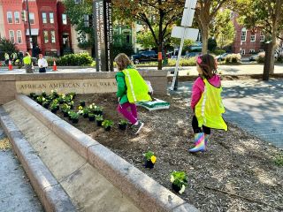 Two young girls (Councilmember Nadeau’s daughters) walk through the dirt patch where they are planting flowers in front of the memorial. “African American Civil War Memorial” is etched into the stone.