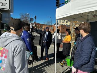 A group gathers under a white tent after a clean-up event. A man wearing a blue winter hat points with his hand off to the left.