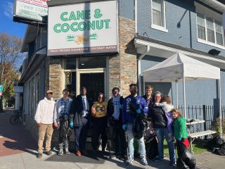 Group poses outside Cane & Coconut storefront wearing gloves and holding trash bags after clean-up event. A white tent is off to the side.