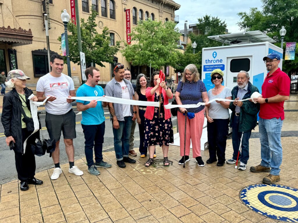 Councilmember Nadeau holds large scissors and cuts toilet paper held by a line of people in front of Throne bathroom in Columbia Heights plaza