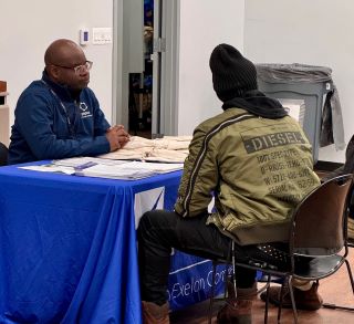 Two men sit at a table across from each other with papers spread on the table