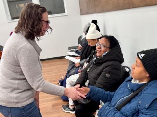 CM Nadeau shakes hands with a woman seated in a folding chair wearing a winter hat as others are seated in a line