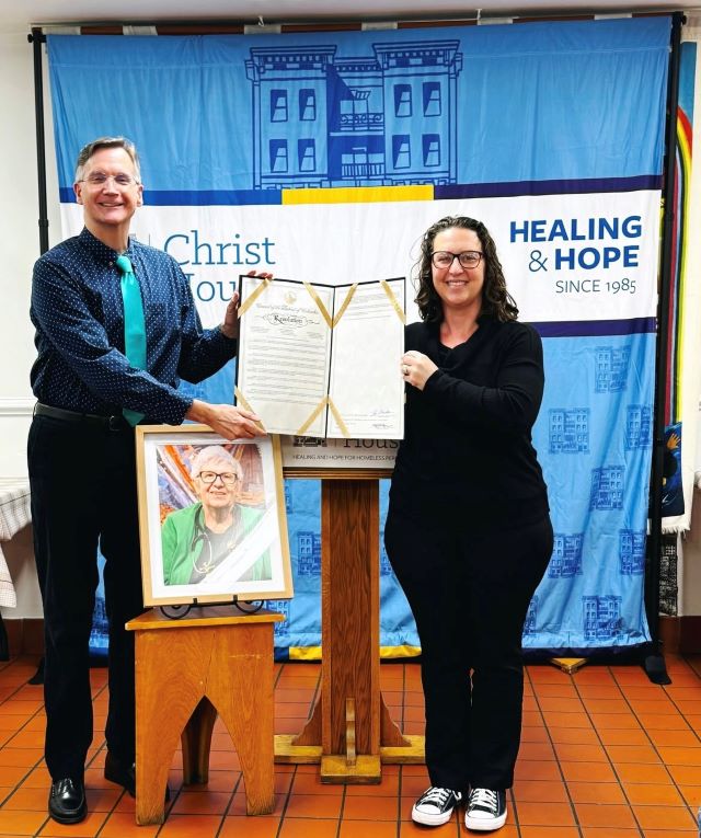 Councilmember Brianne K Nadeau holds a Council resolution with the director of Christ House DC with a framed photo of Dr. Janelle Goetcheus