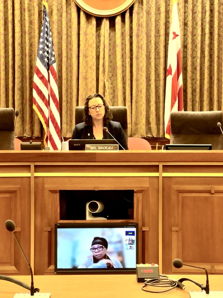 Councilmember Nadeau sits on the dais in the DC Council chamber while a witness appears via Zoom on the screen below. The US flag and DC flag are in the background.