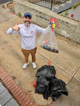 A man poses for a photo with trash bags, he gives a thumbs up and holds up a clear trash bag filled with bottles and cans