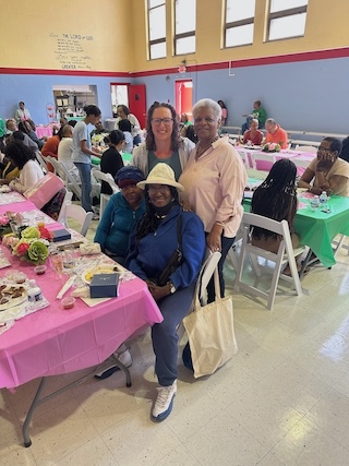 Councilmember Nadeau poses for a photo with three women, two seated and one standing, at a table covered in a pink tablecloth and food and flowers