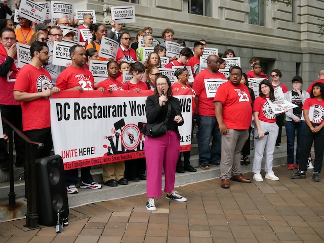 Councilmember Brianne K Nadeau speaks into a microphone in front of a crowd standing on the steps to the Wilson Building and wearing red t-shirts and holding a banner that reads "DC Restaurant Workers" and signs that read "No I82 Repeal Stop the Steal" and "Taxpayer $$$ Good for Jobs"