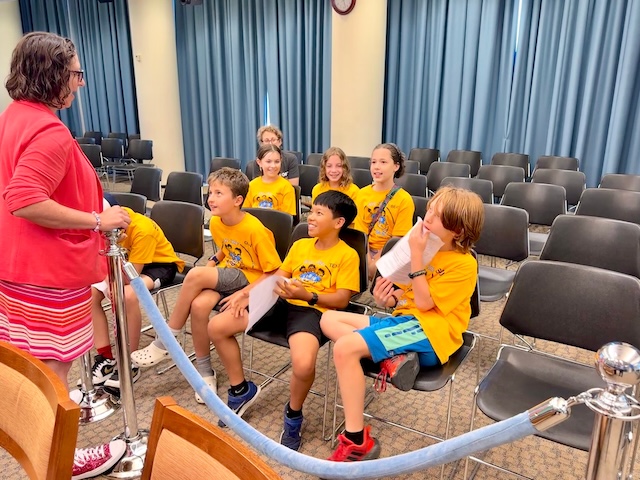 Students wearing yellow shirts are seated in a hearing room and facing Councilmember Nadeau who is answering their questions.