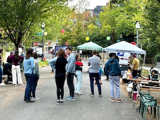 Small groups of people congregate on a street that’s been closed off with pop-up tents and balloons on the background.