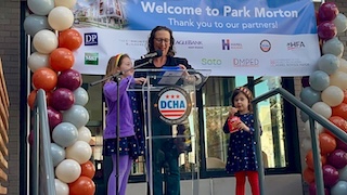 Councilmember Nadeau speaks at a podium with her two young girls by her side. Multicolor balloons are on either side and a "Welcome to Park Morton" sign hangs behind them.
