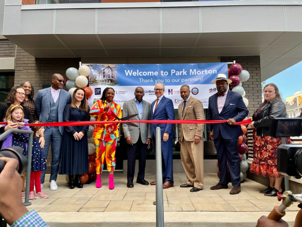 A group of a dozen people, including Councilmember Nadeau, stands out front of a building in front of a long red ribbon with one person holding oversized scissors, ready to cut it.