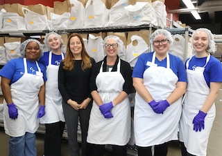 Staff from Councilmember Nadeau's office and Councilmember Brianne K Nadeau, all wearing aprons and hairnets, pose for a photo in front of shelves with grocery bags, with a staff member from Food and Friends
