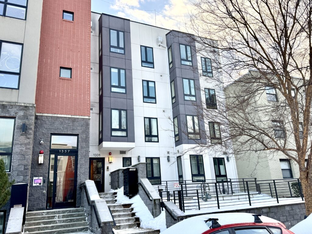 Two small multifamily buildings on a street with fresh snow on the sidewalk.