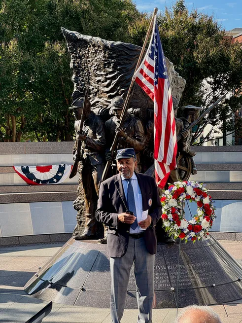 Dr Frank Smith speaks at the African American Civil War Memorial. An American flag and a wreath with red, white, and blue flowers is visible.