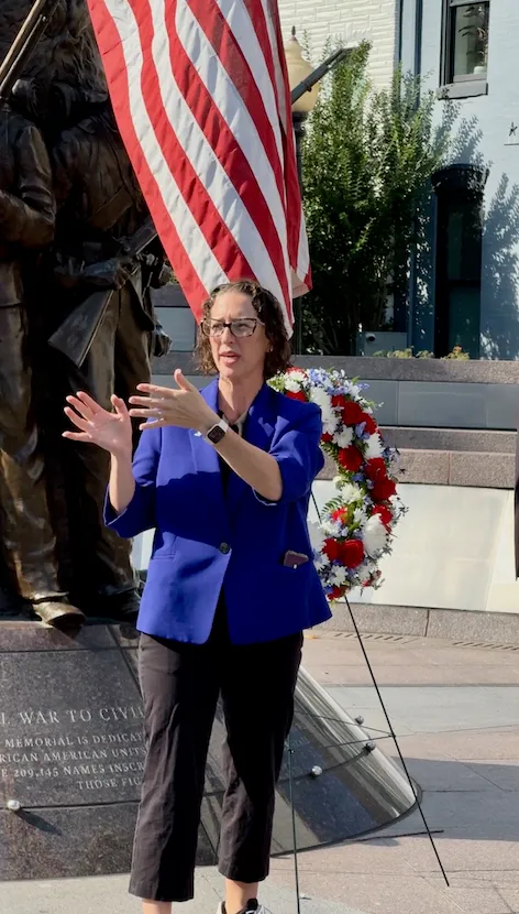 Councilmember Nadeau speaks at the African American Civil War Memorial. An American flag and a wreath with red, white, and blue flowers is visible.